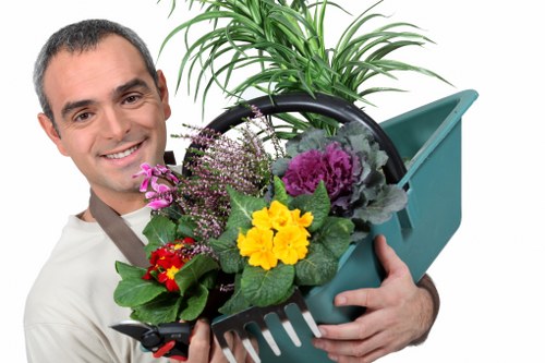 A young man with a friendly expression is holding a green plastic garden toolbox filled with a variety of colorful flowering plants and green foliage, including purple, yellow, and red blooms. The toolbox contains gardening tools, such as a small rake, set against a background of outdoor greenery and a potted plant with lush, long, green leaves. The scene suggests outdoor gardening or planting activities, typical of residential gardens in Enfield, with natural daylight illuminating the vibrant colors of the flowers. The garden area appears well-maintained, with visible healthy plants and a tidy environment, reflecting professional gardening services offered by Garden Maintenance Enfield. The setting emphasizes the attention to detail and care involved in garden landscaping and maintenance, suitable for a front or back garden environment, highlighting the importance of flowering plants, soil preparation, and outdoor aesthetics related to gardening.