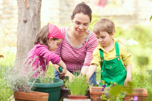 A woman with dark hair tied back, wearing a pink and white striped long-sleeve shirt, is engaged in gardening activities outdoors in a residential garden in Enfield. She is accompanied by a young girl in a bright pink headscarf and matching top, and a young boy in a yellow shirt with green overalls. They are tending to various potted plants placed on a garden table composed of terracotta and plastic containers, featuring herbs and flowering plants. The garden setting includes a lush green lawn, mature trees, and a wooden fence in the background, with sunlight filtering through the leaves, indicating a clear day. The scene emphasizes outdoor family gardening activities, with the children actively involved in planting or watering under the woman’s supervision. The environment is well-maintained, demonstrating typical garden maintenance and landscaping efforts common in Enfield. This image visually supports services related to gardening and lawn care, illustrating engaging garden work suitable for residential outdoor spaces in the local area.