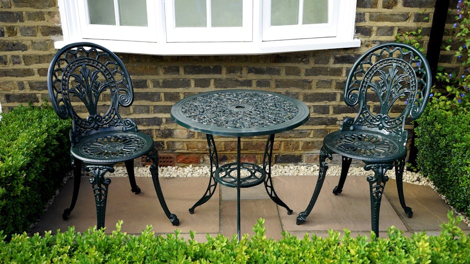 A small outdoor garden seating area featuring a round, ornate metal table with an intricate floral pattern on the top, flanked by two matching wrought iron chairs with decorative backrests and curved