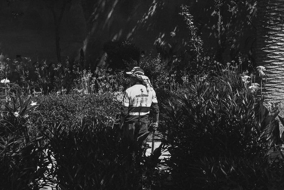 A gardener wearing a wide-brimmed hat and a high-visibility jacket marked with 'SOMETHING' is working at night in a garden bed filled with dense, leafy plants. The scene is illuminated by an external