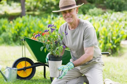 A smiling elderly man wearing a straw hat, grey t-shirt, and gardening gloves crouches on a well-maintained lawn in a backyard garden. He is holding a pot containing flowering plants with purple, red, and pink blooms, which appears freshly planted or prepared for planting. In the background, there is a lush hedge or shrubbery forming a natural boundary, with mature trees providing shade and greenery. To the left, a green wheelbarrow with black handles and yellow wheels is positioned on the grass, partially filled with gardening tools and supplies. The garden features a mix of neatly trimmed grass and a paved pathway, with vibrant natural tones in the foliage and flowers. The weather looks bright and clear, suggesting a pleasant day suitable for outdoor gardening. This scene exemplifies routine outdoor maintenance and planting activities, aligning with professional gardening services offered by companies like Garden Maintenance Enfield for homes in the Enfield area, near postcode EN1.